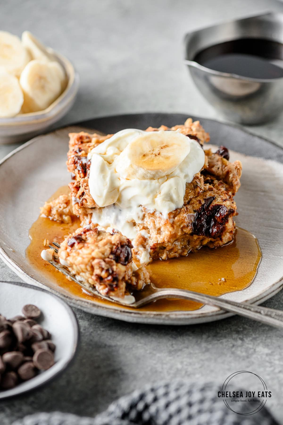 Banana bread baked oatmeal with Greek yogurt, a banana slice, and maple syrup on a grey plate with a bite taken out with a fork.