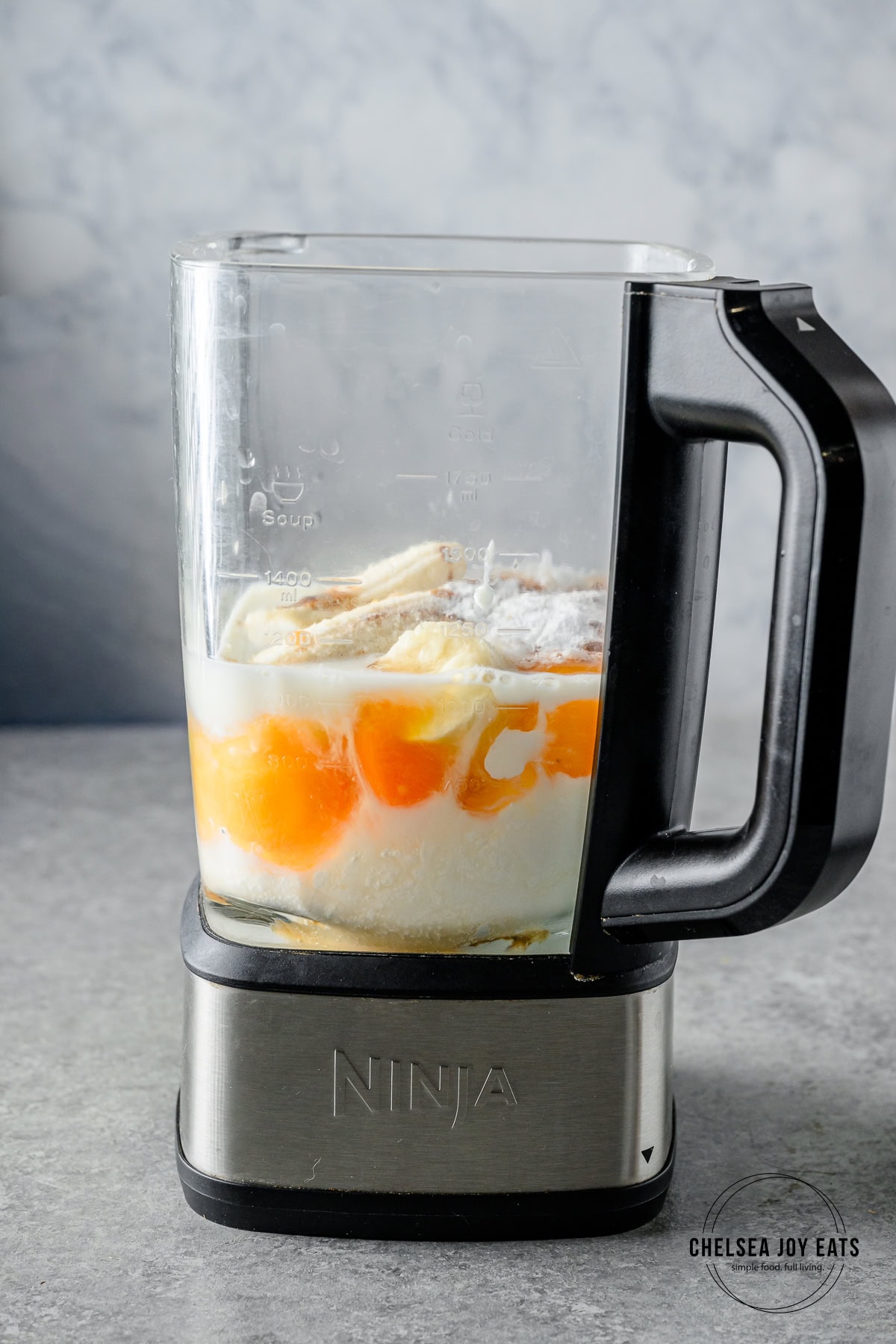 Eggs, bananas, Greek yogurt, and milk in the carafe of a blender waiting to be blended.