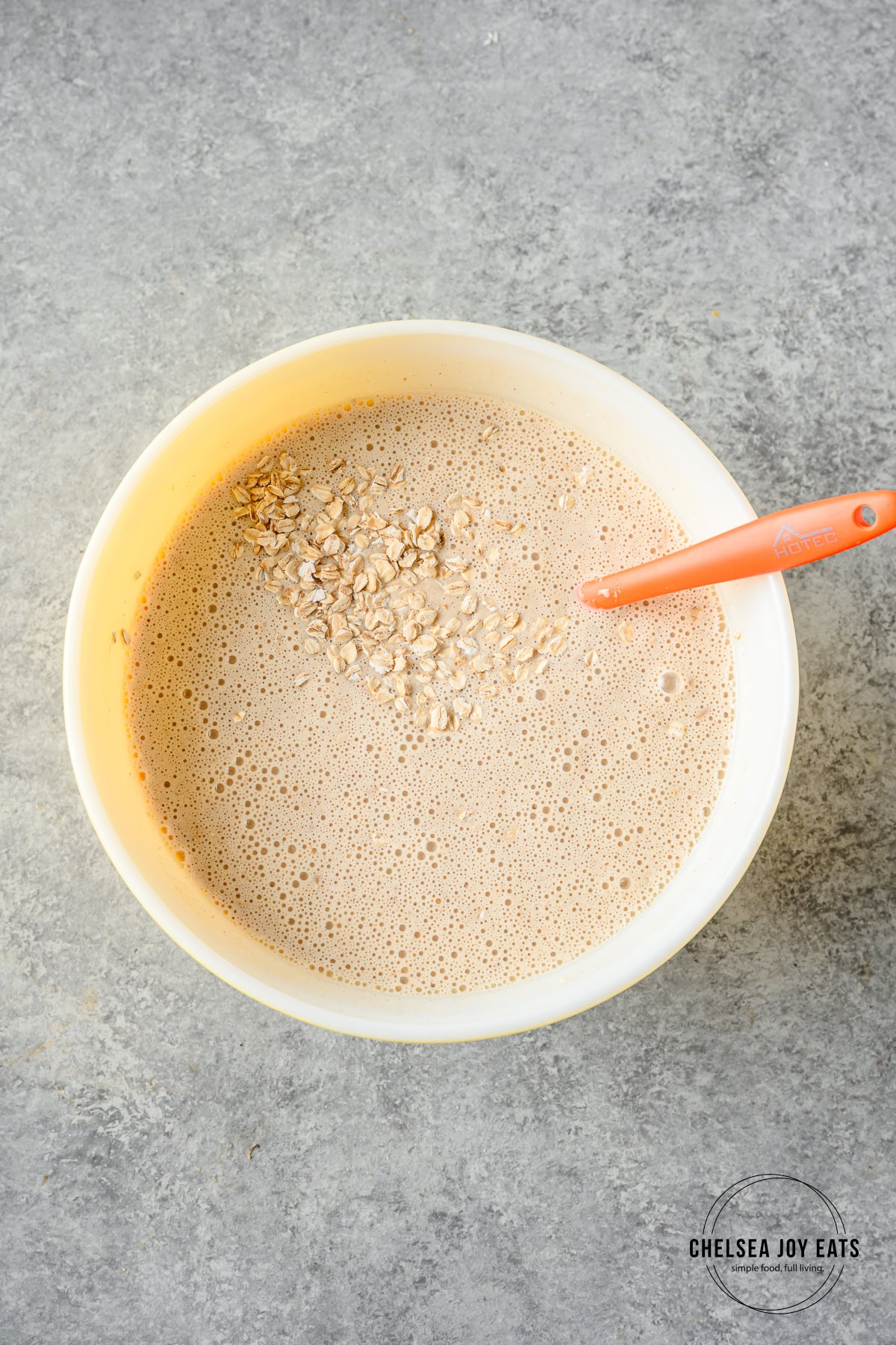 Blended banana and Greek yogurt mixture stirred with gluten free oats in a large ceramic bowl with an orange silicon spatula.
