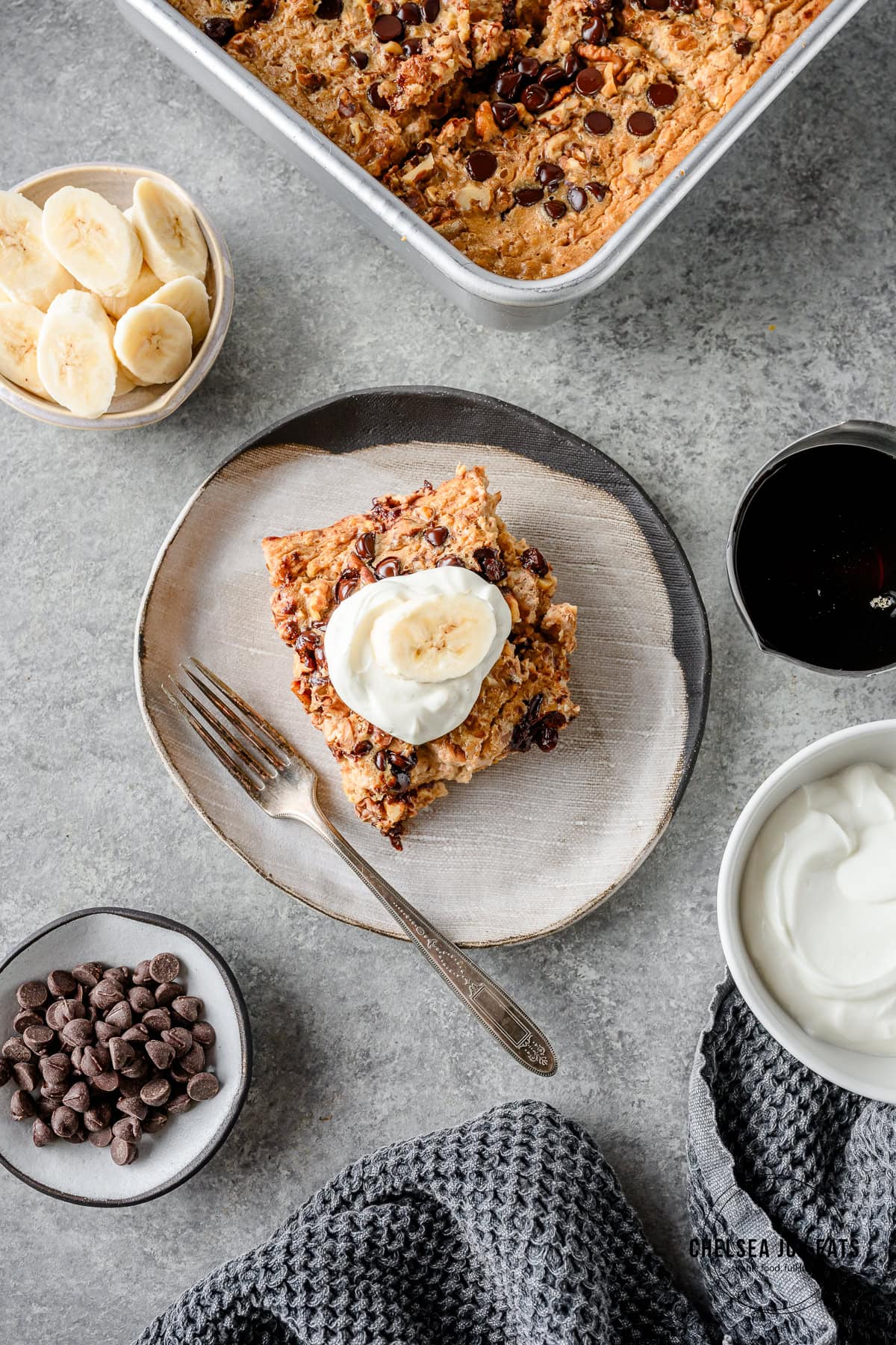 Serving of gluten free banana bread baked oatmeal in a grey plate with a fork with a small dish of fresh bananas, maple syrup, and chocolate chips nearby.