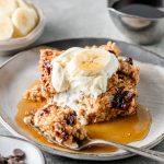 Banana bread baked oatmeal with Greek yogurt, a banana slice, and maple syrup on a grey plate with a bite taken out with a fork.