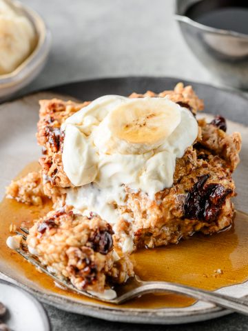 Banana bread baked oatmeal with Greek yogurt, a banana slice, and maple syrup on a grey plate with a bite taken out with a fork.