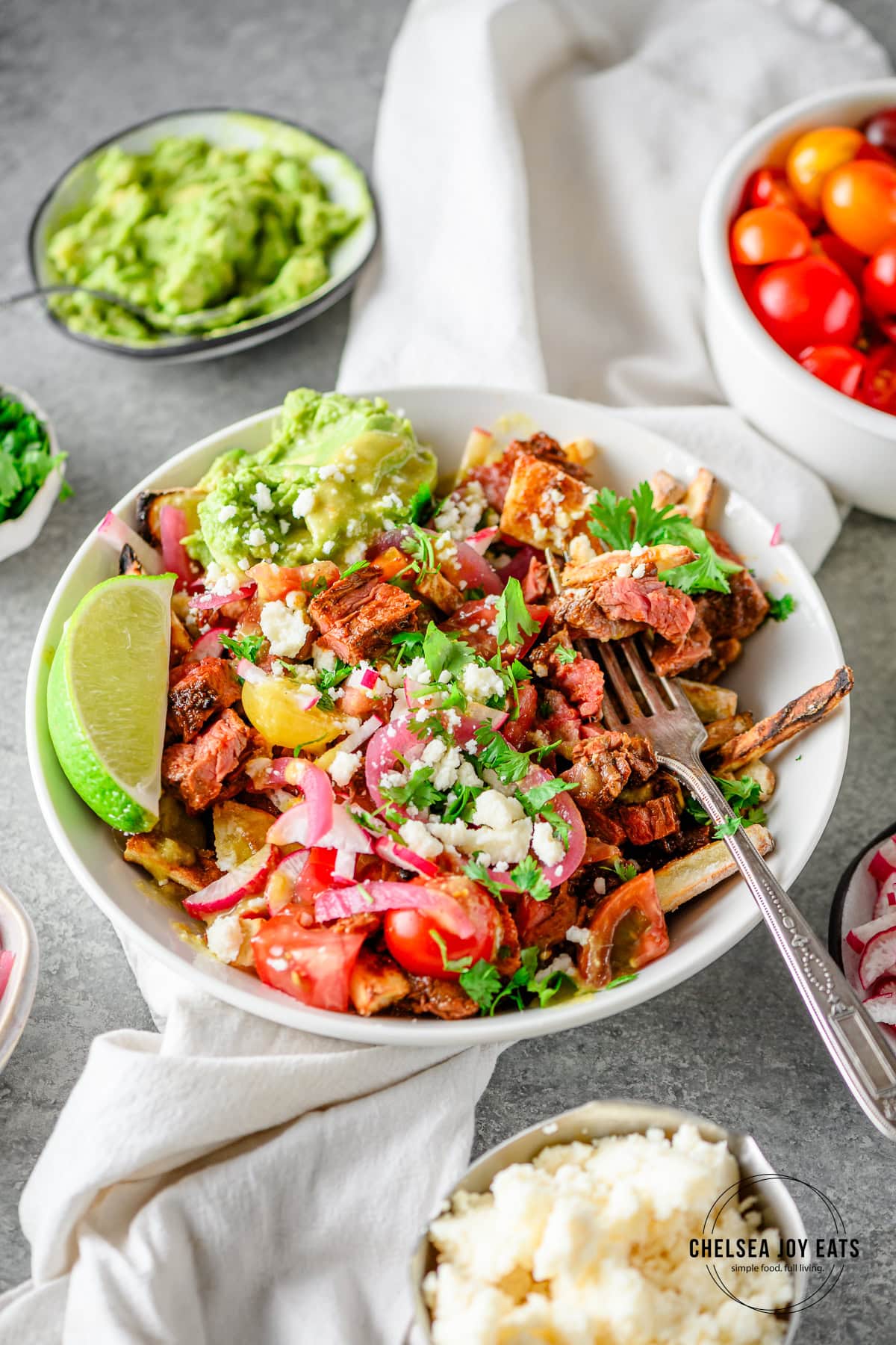Gluten free carne asada fries with sweet potato fries topped with fresh vegetables, guacamole, cilantro, and cotija cheese with extra bowls of toppings alongside.