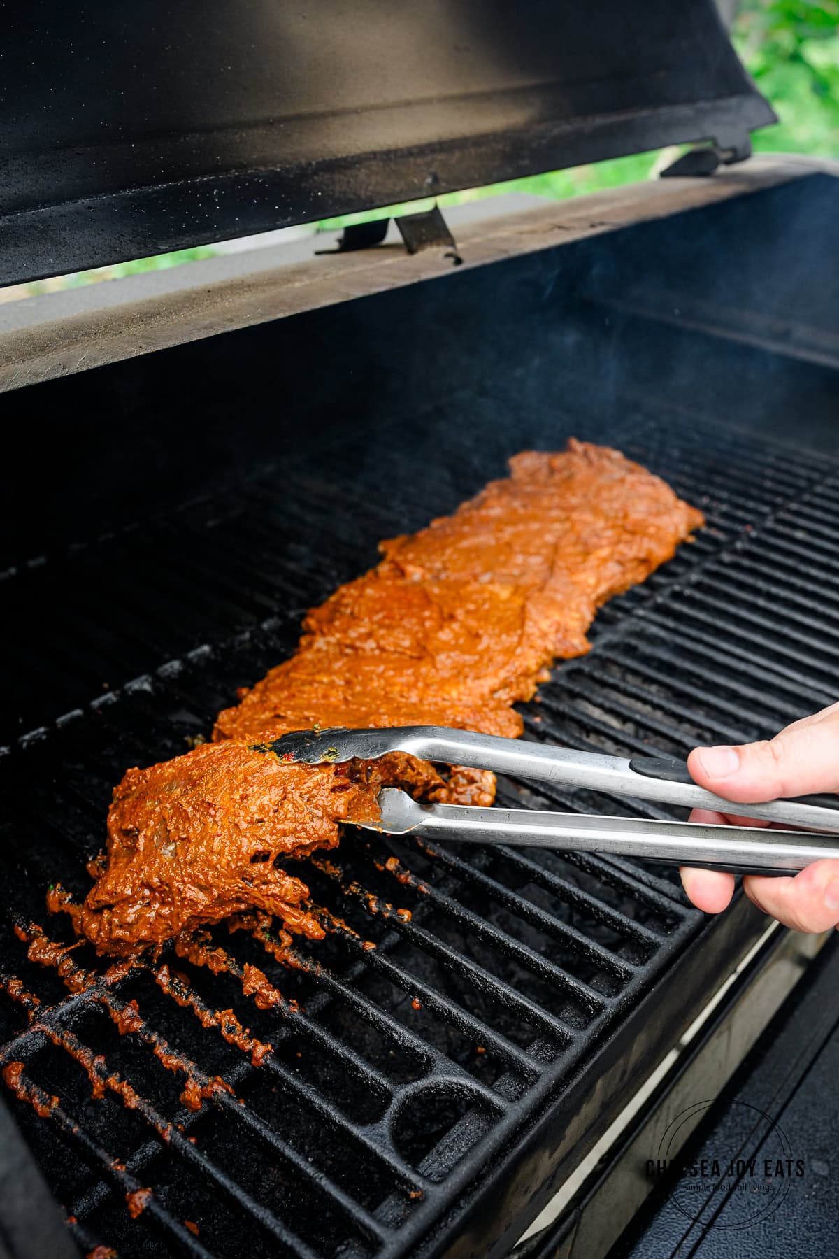 Carne asada steak being flipped on a grill with metal kitchen tongs.