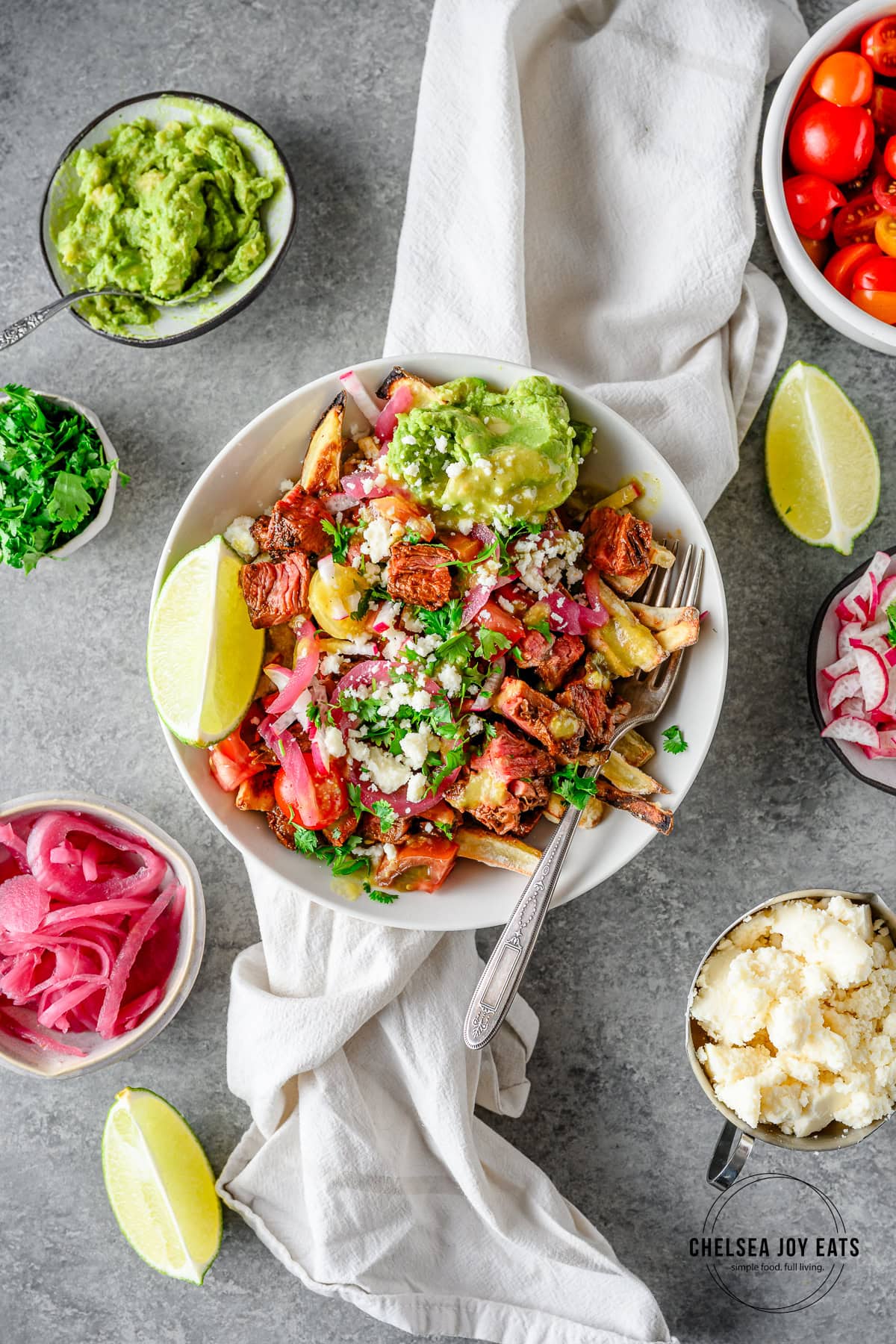 Assembled carne asada sweet potato fries with toppings like guacamole, pico de gallo, pickled red onions, cilantro, and a lime wedge.