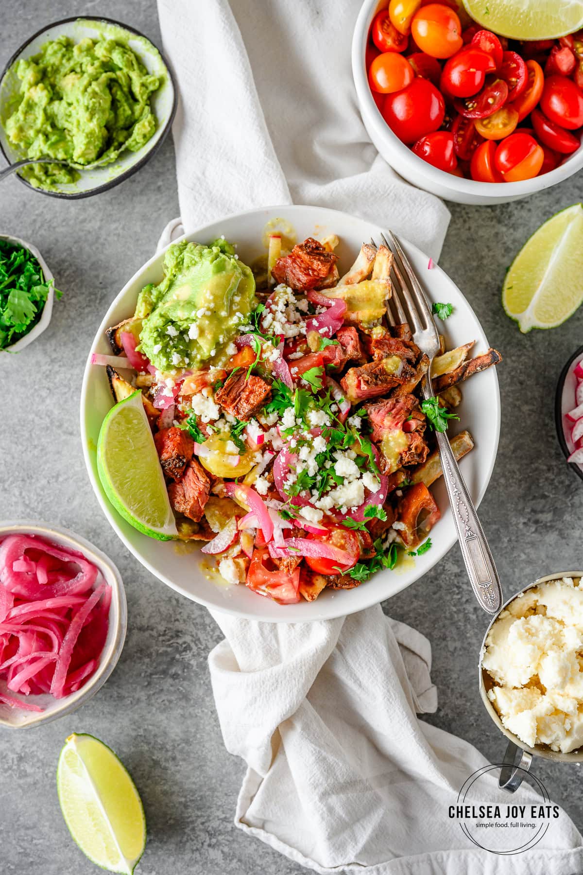 Bowl with a heaping serving of homemade gluten free carne asada fries with bowls of toppings like pickled red onions, cotija cheese, and guacamole surrounding.
