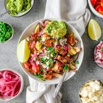 Bowl with sweet potato fries topped with homemade carne asada, guacamole, pickled onions, pico de gallo, and other toppings with extra toppings in bowls.