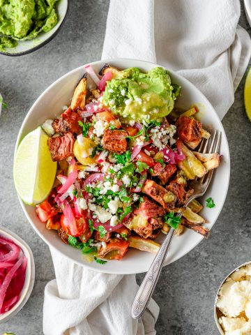 Bowl with sweet potato fries topped with homemade carne asada, guacamole, pickled onions, pico de gallo, and other toppings with extra toppings in bowls.