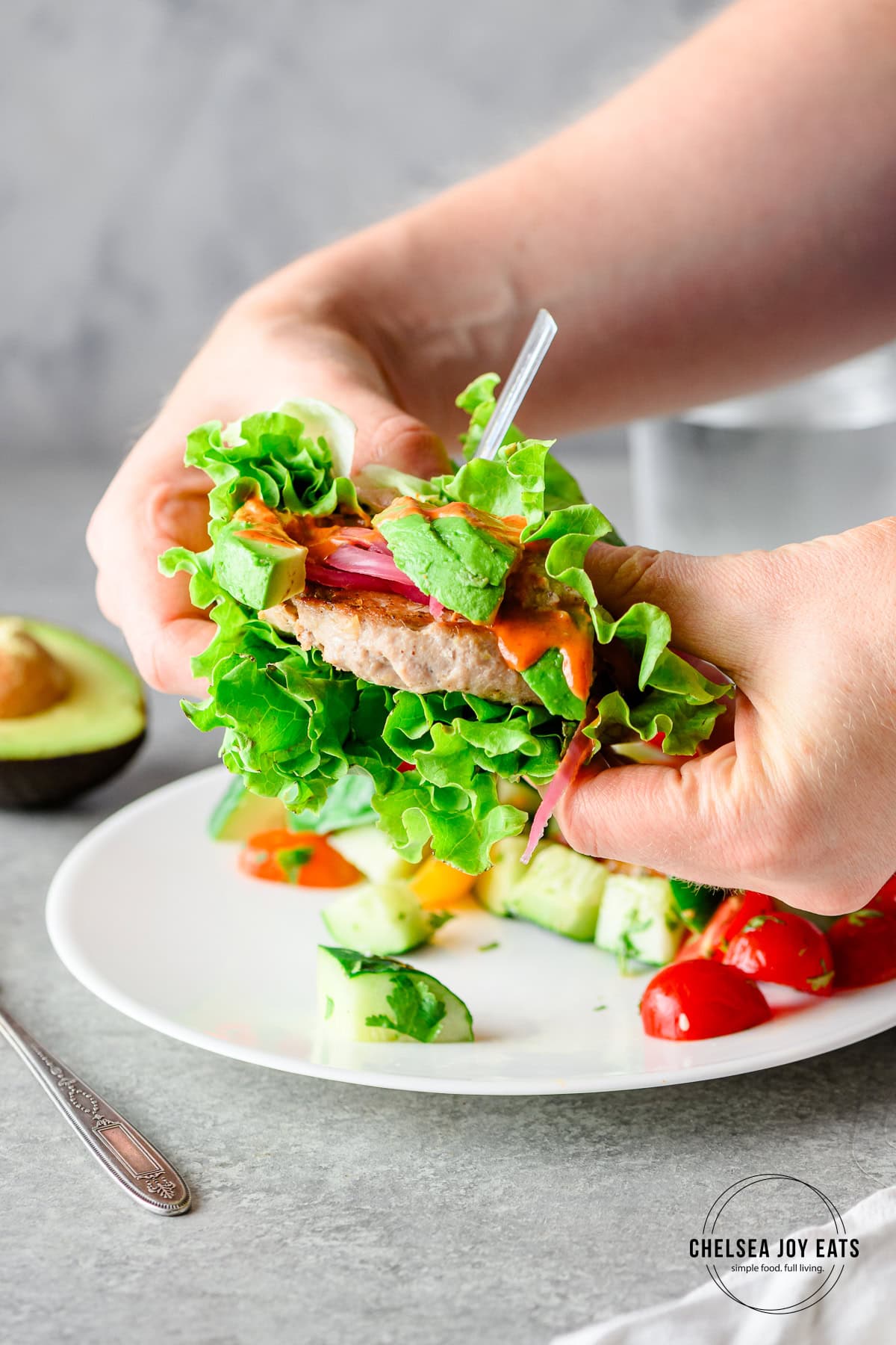 Person holding a lettuce-wrapped tuna burger with avocado, pickled onions, and sriracha mayo.