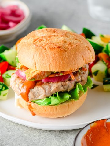 Homemade tuna burgers with pickles onions and sriracha mayo on a gluten free bun served on a plate with a salad.
