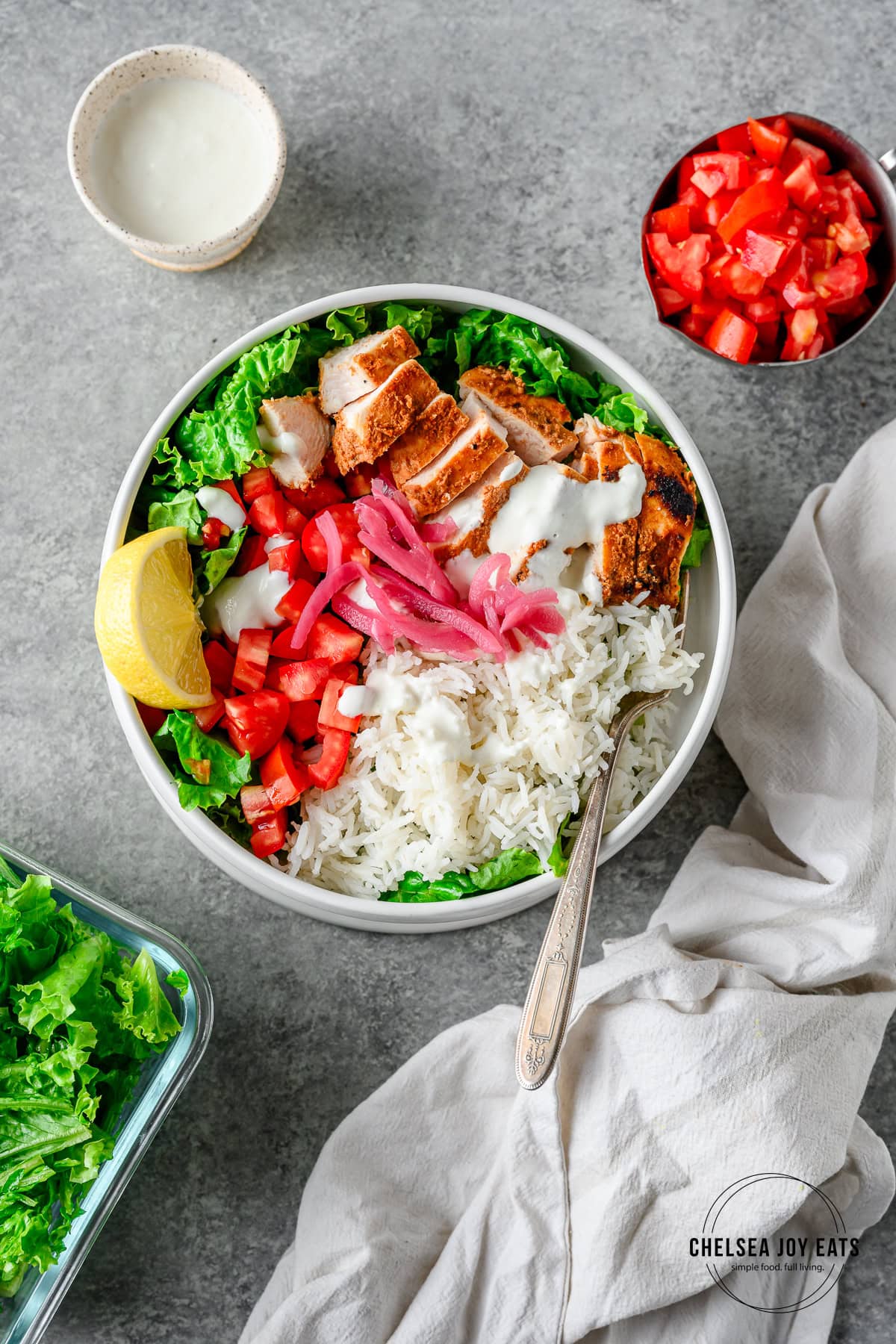 Assembled chicken shawarma bowls with rice, fresh vegetables, sliced chicken shawarma, garlic sauce, and pickled red onions, with more toppings in bowls around.