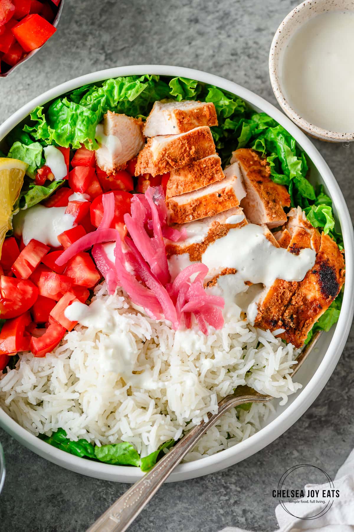 White ceramic bowl with lettuce, rice, tomato, pickled onion, homemade gluten free chicken shawarma drizzled with garlic yogurt sauce served with a fork.