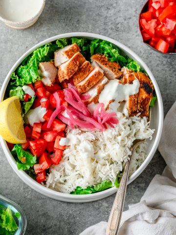 Gluten free chicken shawarama bowls with sliced shawarma chicken breast, green lettuce, rice, tomatoes, pickled onions, and garlic sauce in a white ceramic bowl.