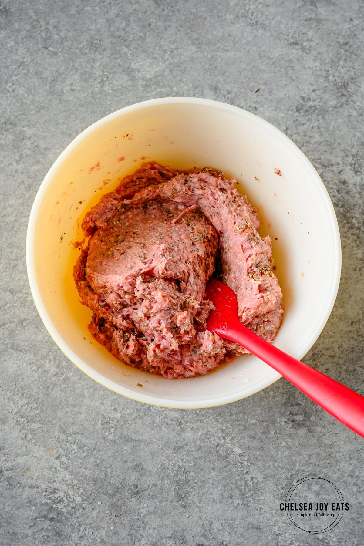 Raw ground turkey mixed with Italian seasoning in a ceramic bowl with a silicon spatula.