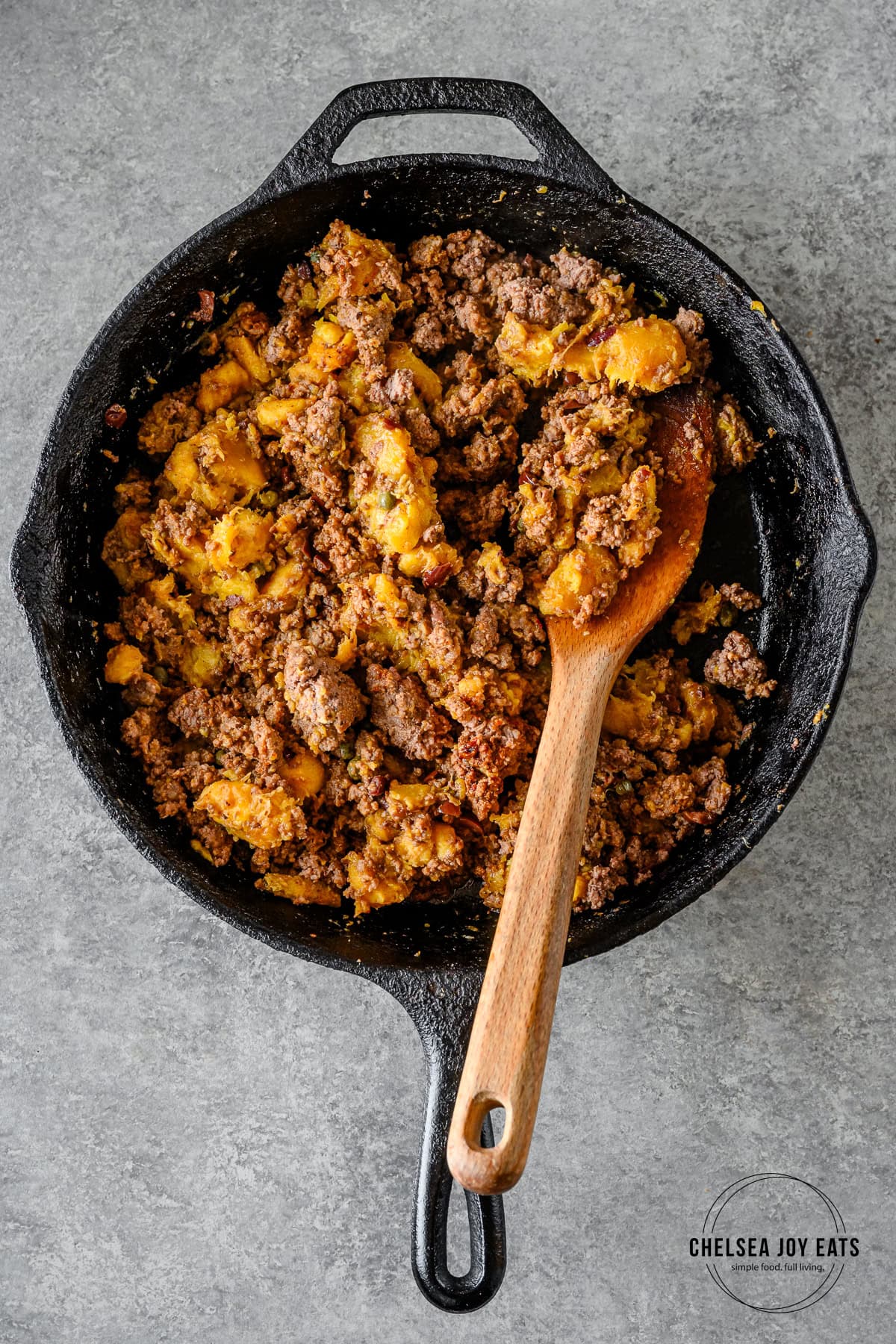 Browned ground beef, mashed plantains, olives, and capers in a cast iron skillet stirred by a wooden spatula.