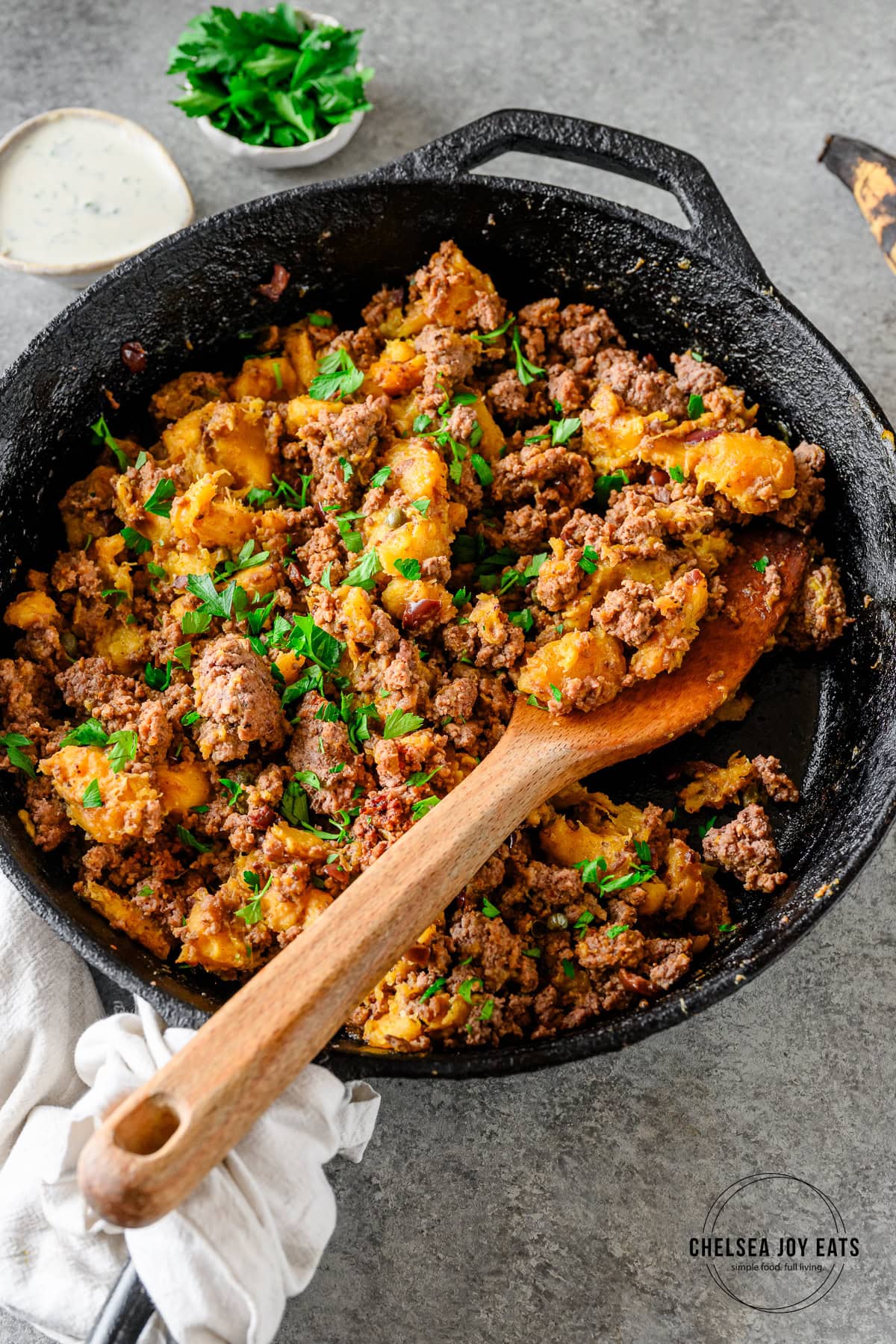 Plantain and ground beef mixture with olives and capers in a cast iron skillet being scooped by a wooden spatula.