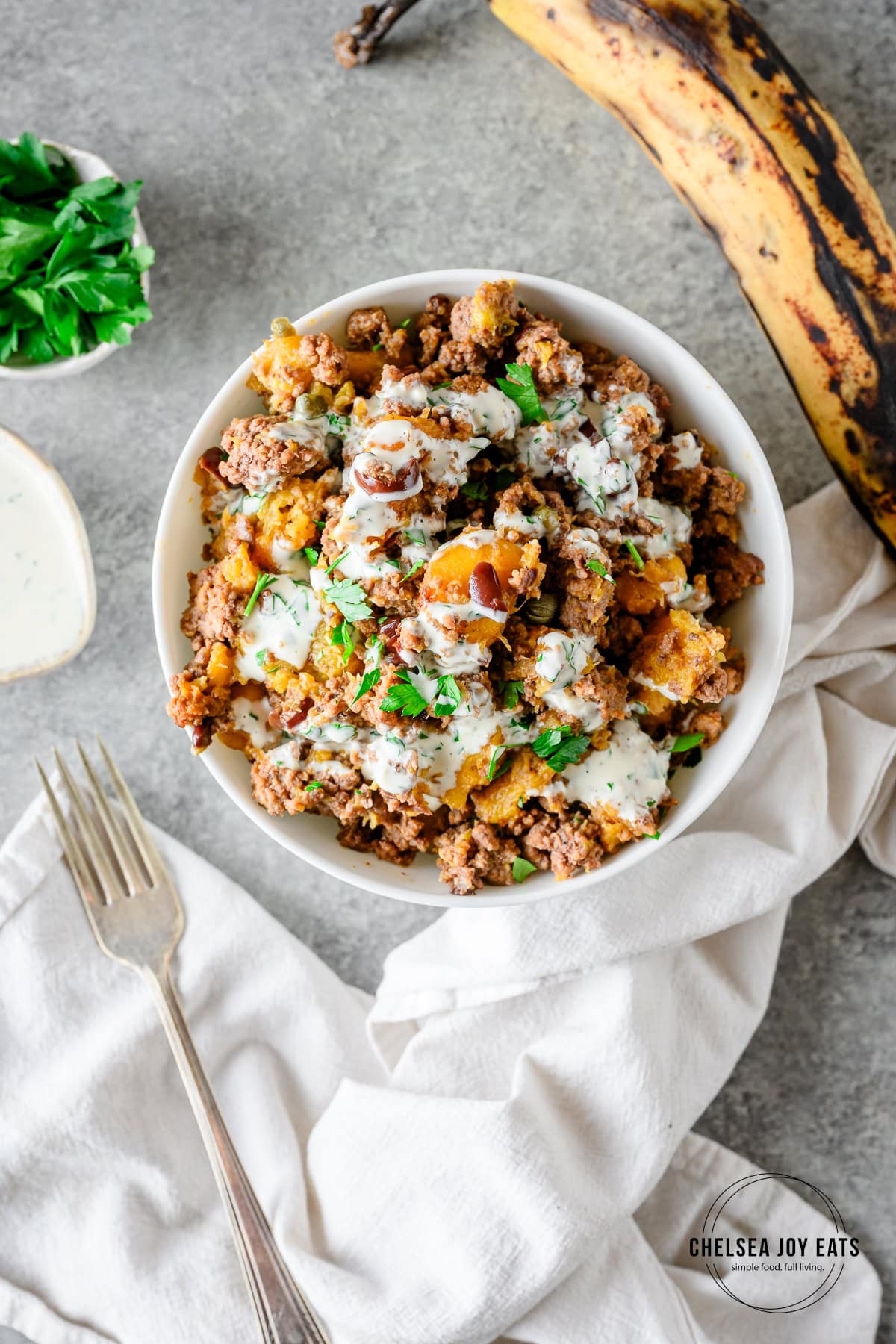 Plantain ground beef bowl with olives and capers, topped with mayo sauce and served in a white bowl on a grey countertop with linens, parsley, and plantains alongside.