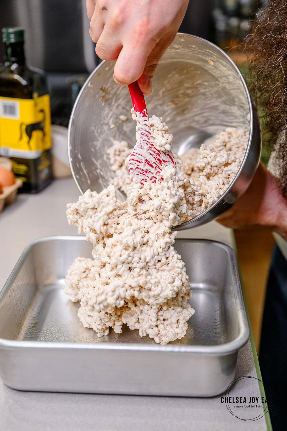 Person dumping marshmallow-coated crispy rice cereal into a greased 8x8 baking pan.