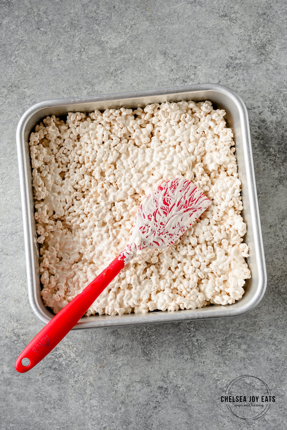 Rice krispies mixture spread in an 8x8 baking pan with a silicon spatula on a light background.