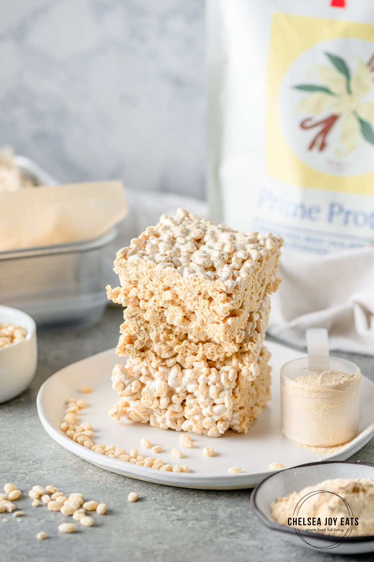 Homemade rice krispies treats stacked on a plate with a scoop of protein powder besides.