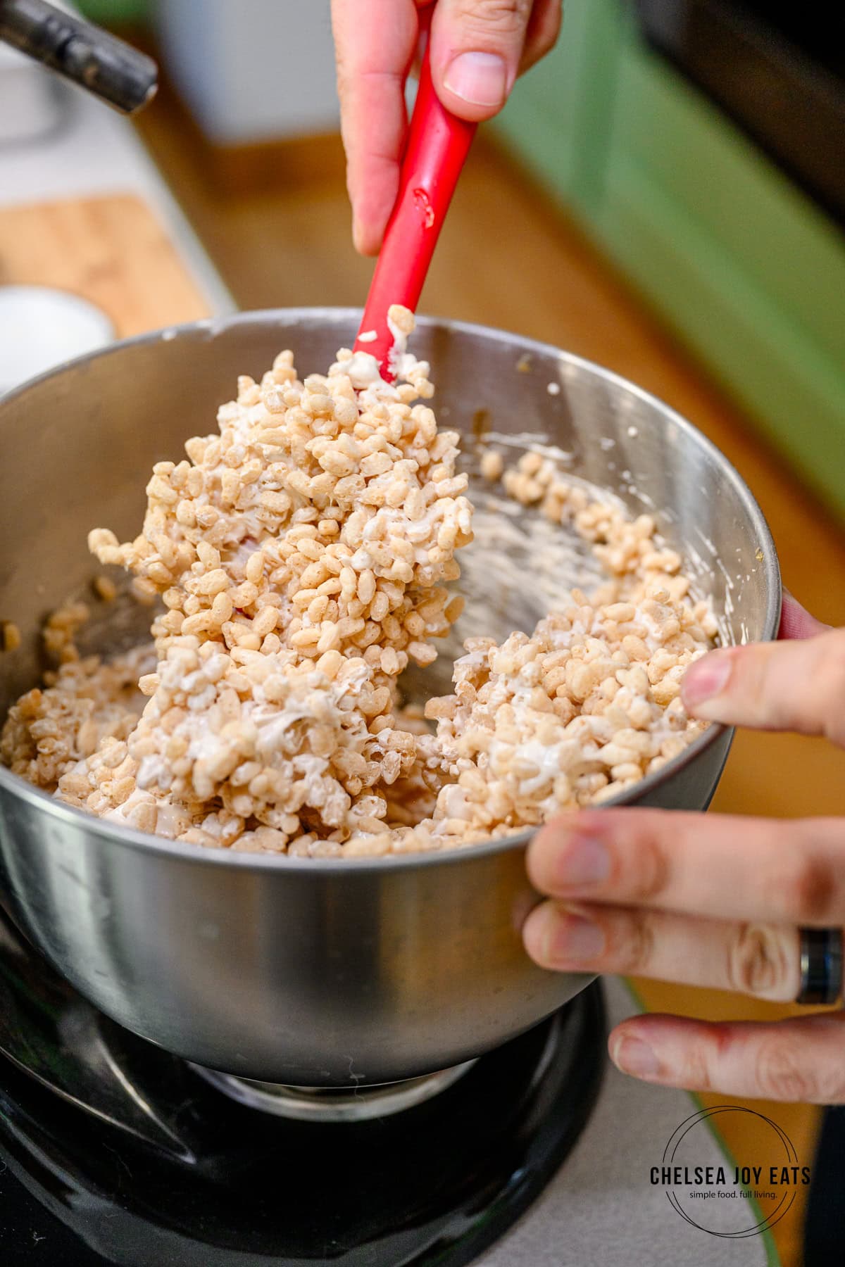 Person stirring crispy rice cereal into a marshmallow mixture in the bowl of a stand mixer with a silicon spatula.