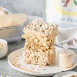 Homemade protein rice krispies bars stacked on a white plate with crispy rice cereal and a scoop of protein powder besides.