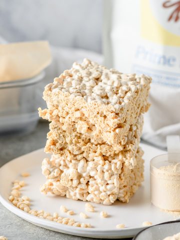 Homemade protein rice krispies bars stacked on a white plate with crispy rice cereal and a scoop of protein powder besides.