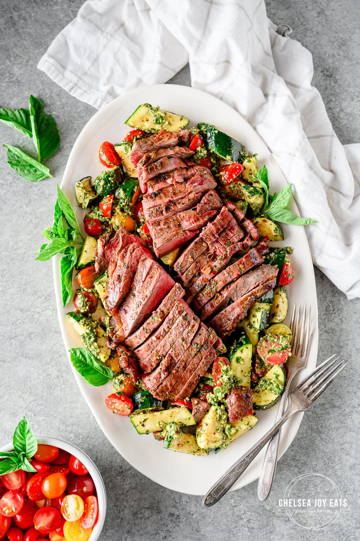 Grilled flank steak salad served on a platter, with a grilled zucchini and tomato salad mixed with fresh pesto and topped with sliced grilled flank steak.