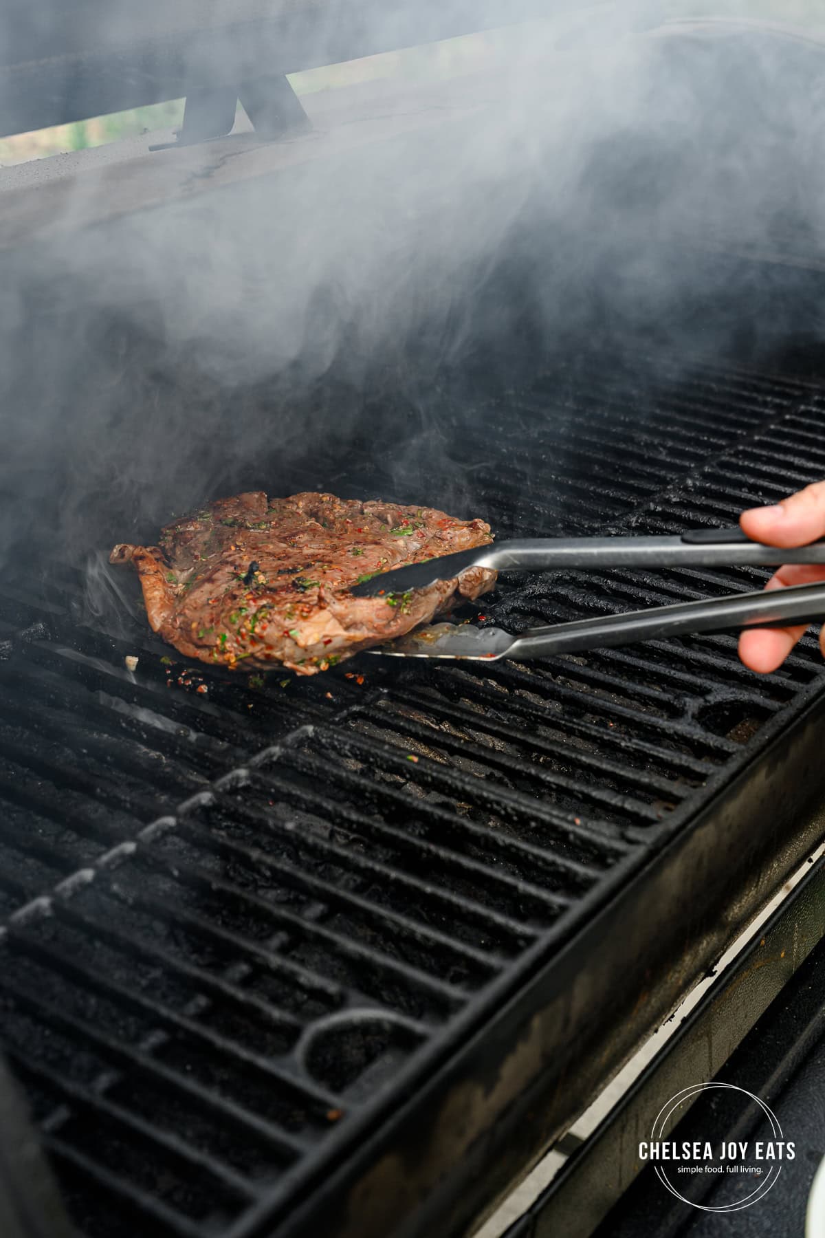 Marinated flank steak being flipped on a grill with metal kitchen tongs.
