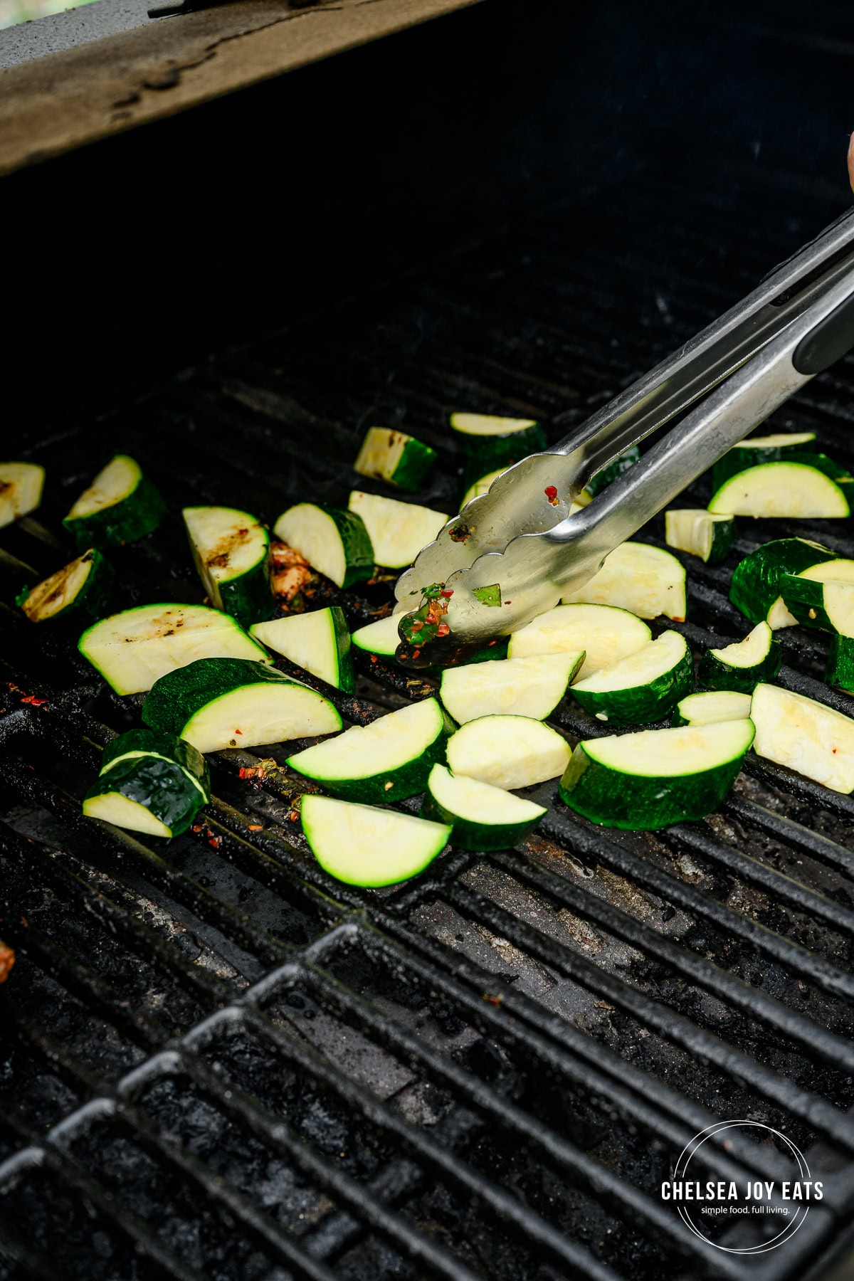 Zucchini slices grilling on a grill and being flipped with metal kitchen tongs.