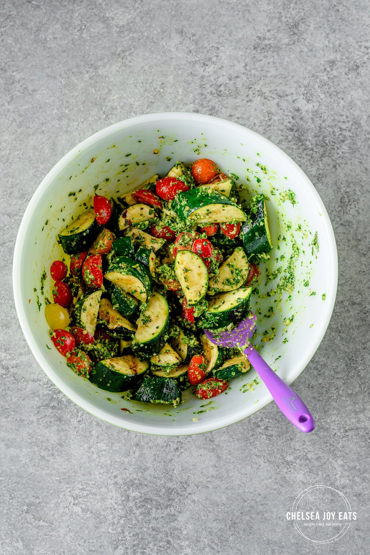 Zucchini and tomatoes stirred with pesto in a large ceramic bowl with a silicone spatula.
