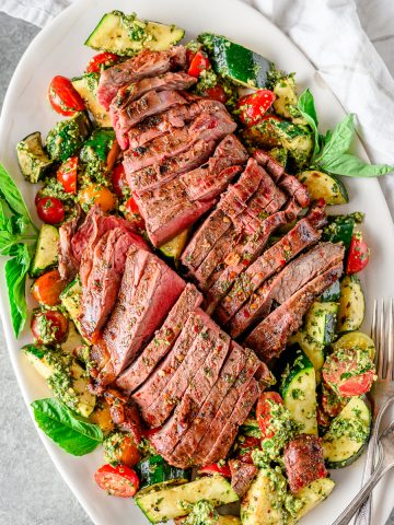 Grilled flank steak salad served on a platter, with a grilled zucchini and tomato salad mixed with fresh pesto and topped with sliced grilled flank steak.
