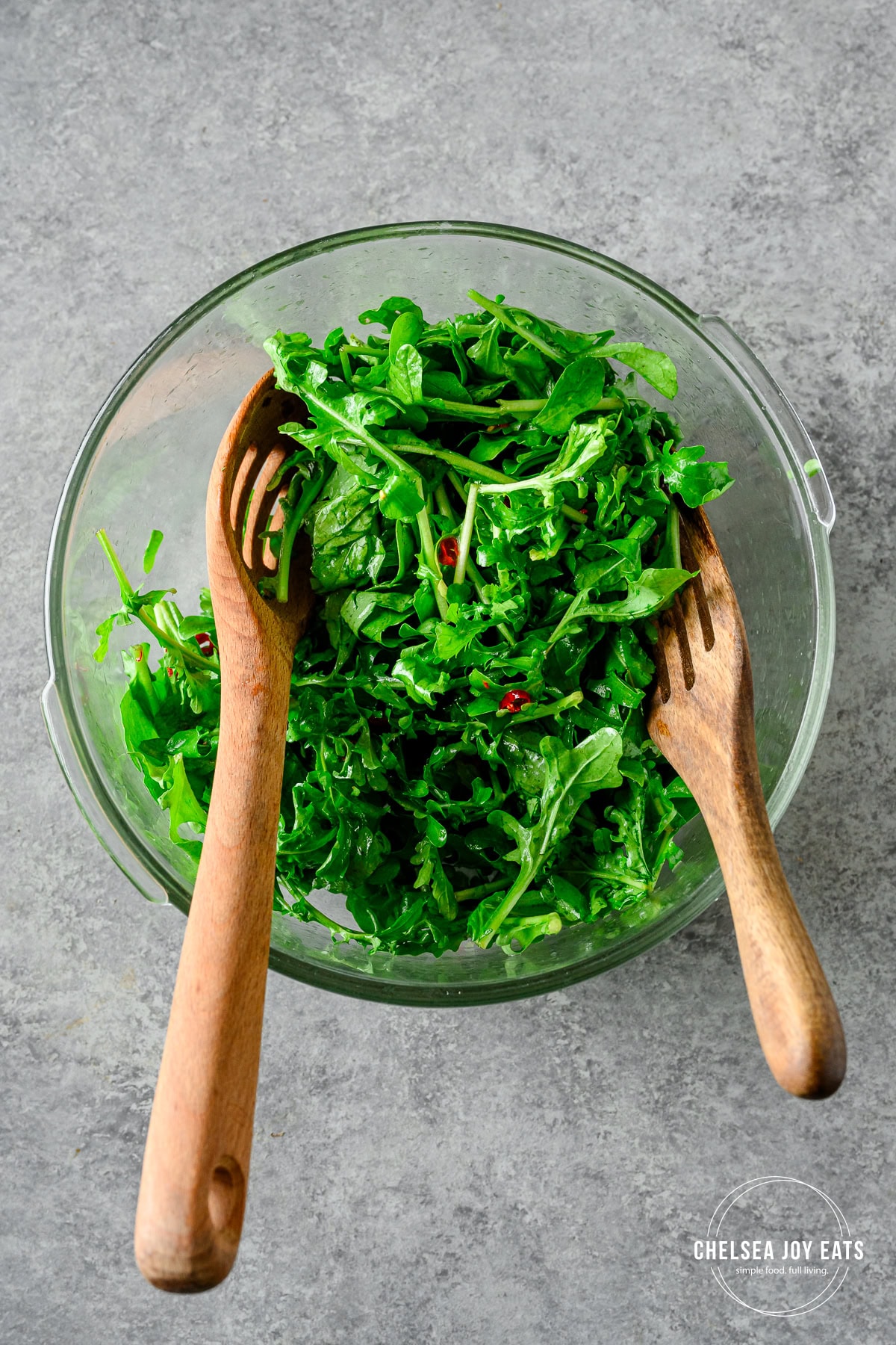 Fresh arugula tossed with chili honey lime dressing with two wooden spoons in a large glass bowl.