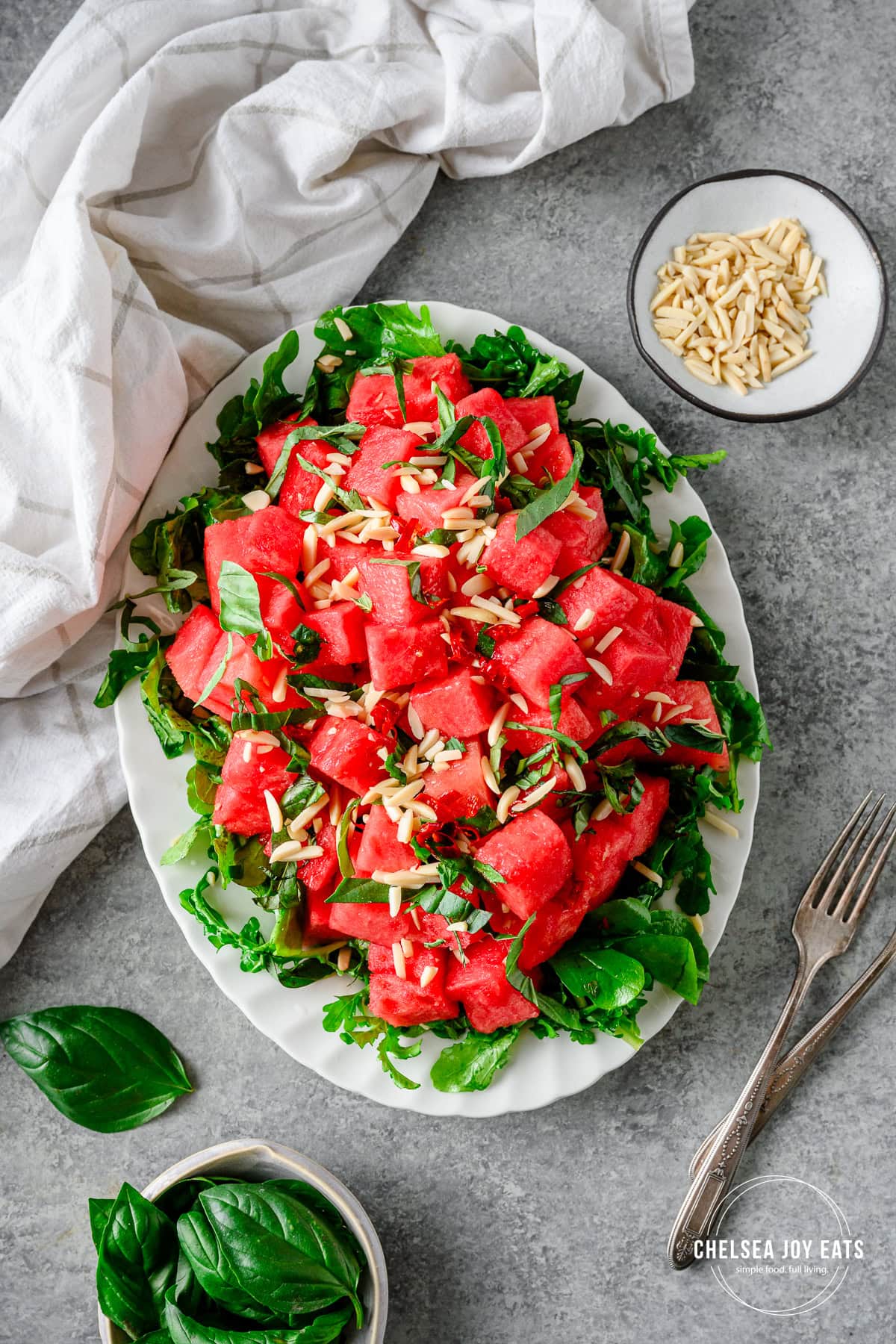 Fresh arugula and watermelon salad sprinkled with slivered almonds and fresh herbs on a white platter.