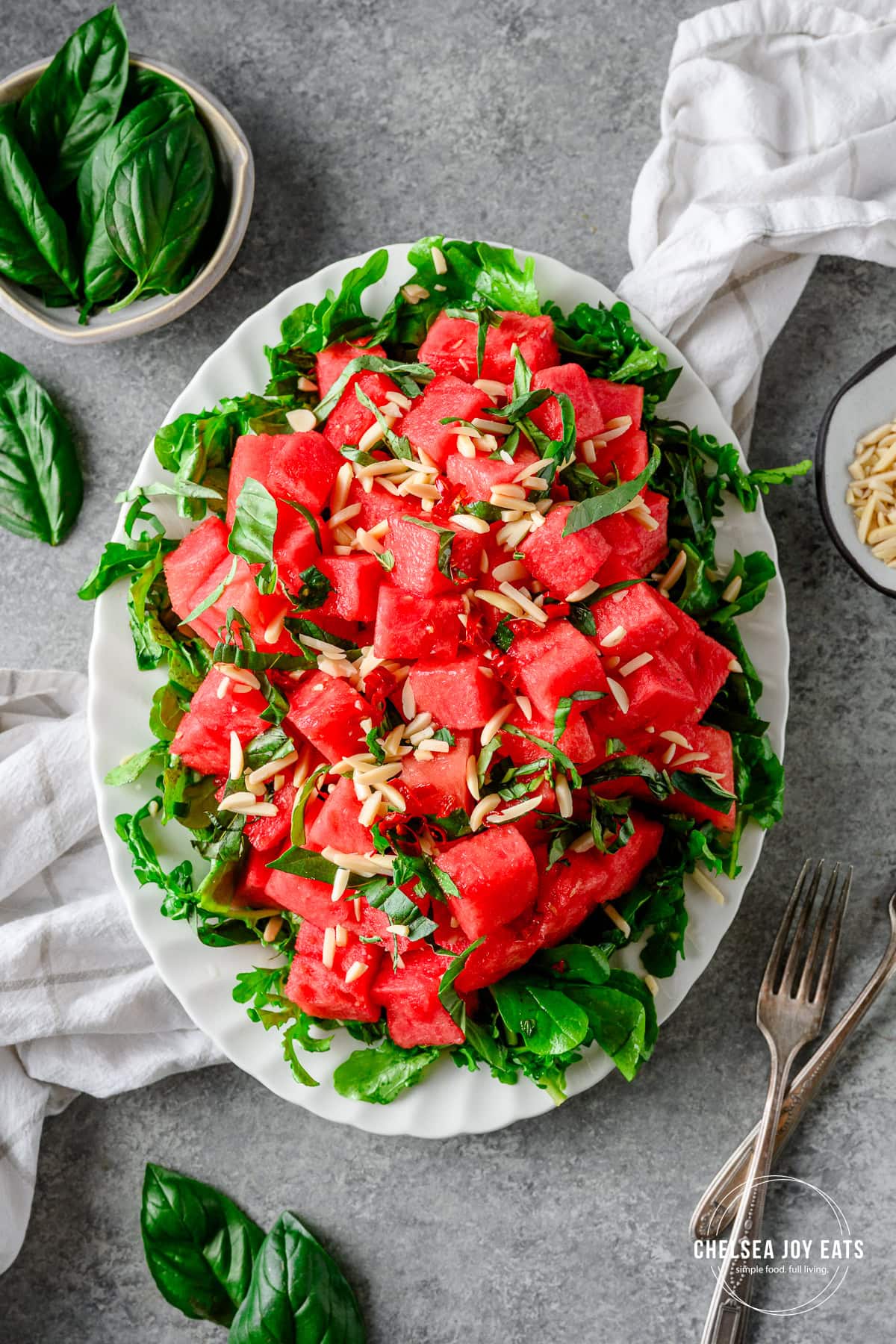 Watermelon Arugula Salad with slivered almonds and drizzled with honey lime dressing on a white platter with fresh basil besides.