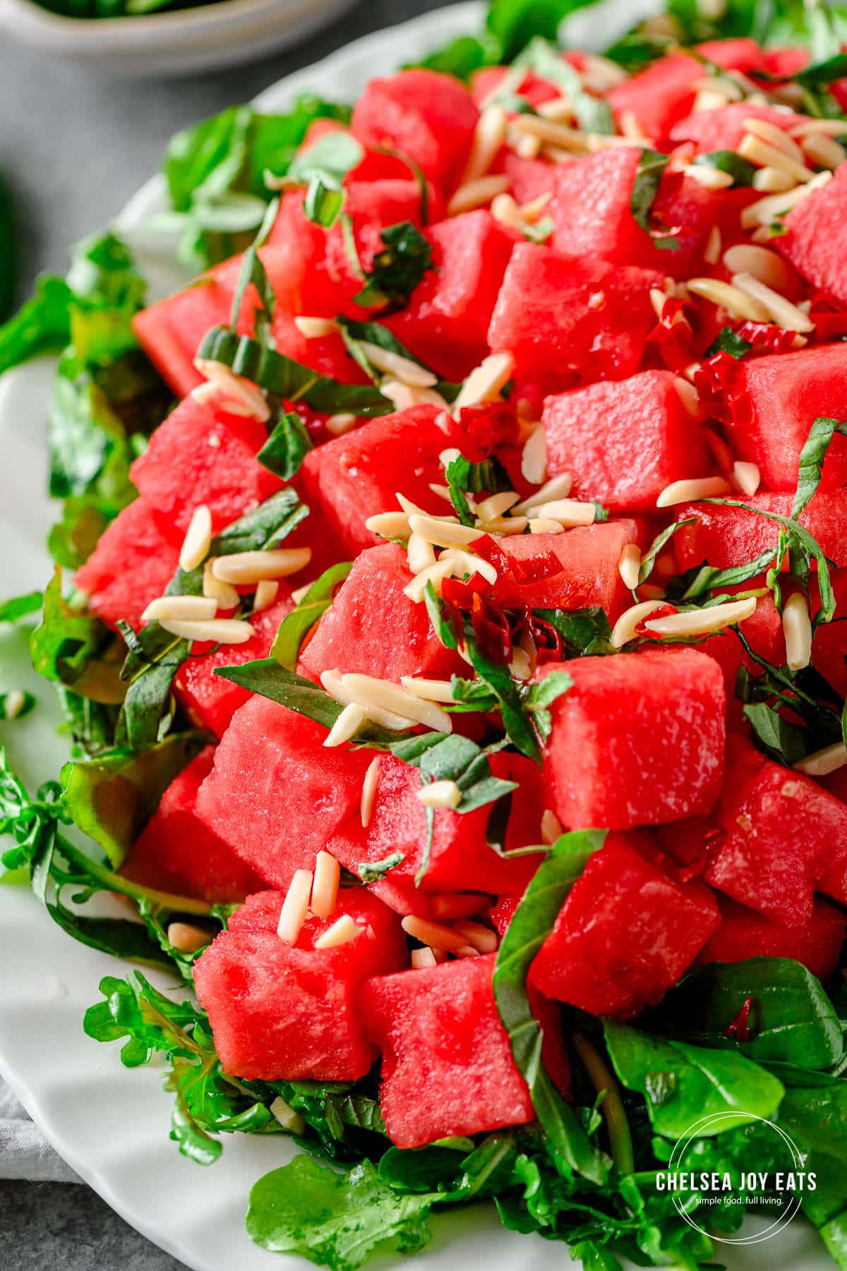 Watermelon served over arugula and topped with slivered almonds, mint, basil, and chili honey lime dressing on a white platter.