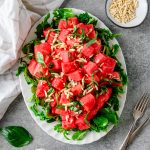 Watermelon Arugula Salad with slivered almonds and drizzled with honey lime dressing on a white platter with fresh basil besides.