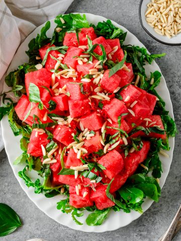 Watermelon Arugula Salad with slivered almonds and drizzled with honey lime dressing on a white platter with fresh basil besides.
