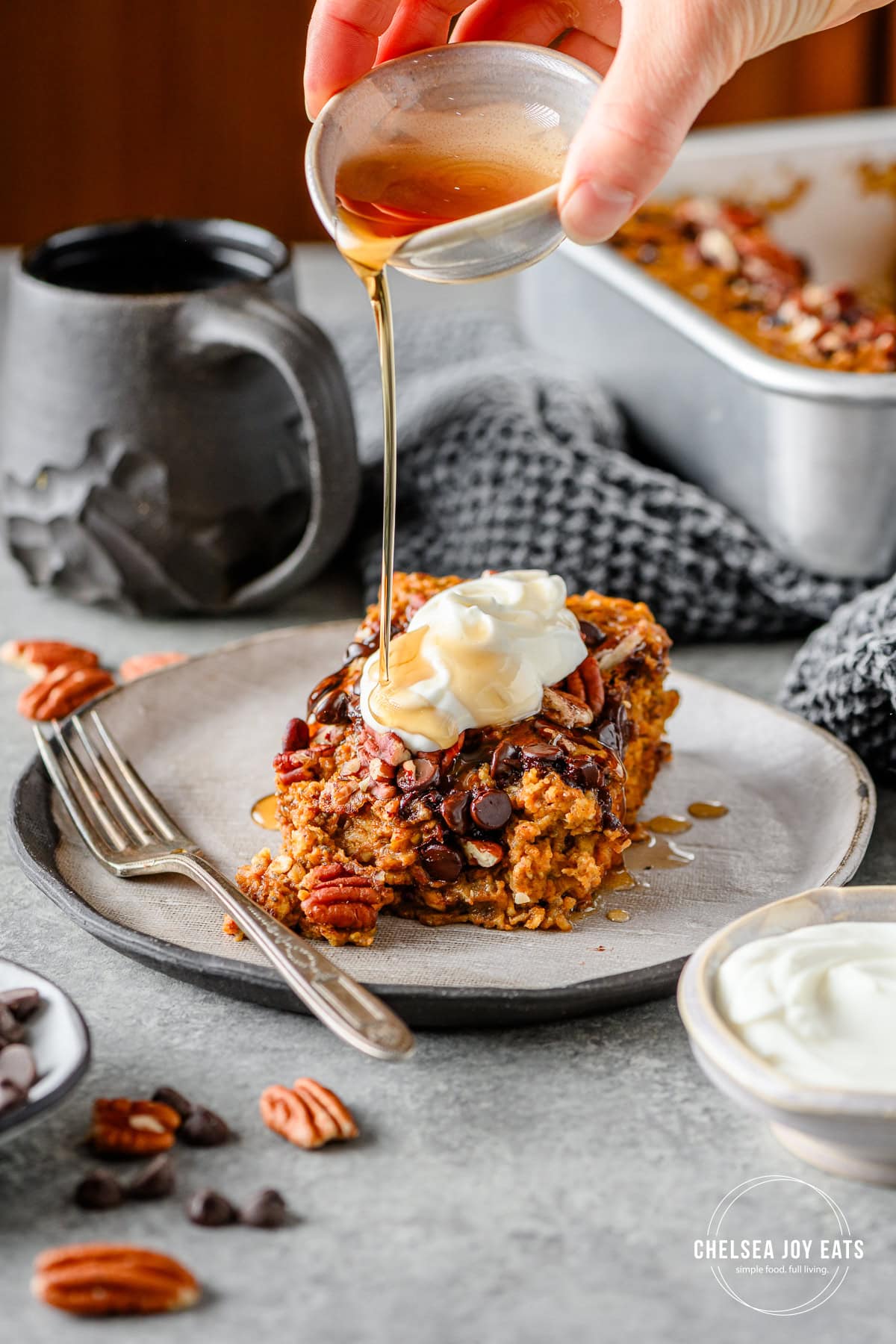 Serving of gluten free pumpkin baked oatmeal topped with Greek yogurt and drizzled with maple syrup on a ceramic plate with a cup of coffee in the background.