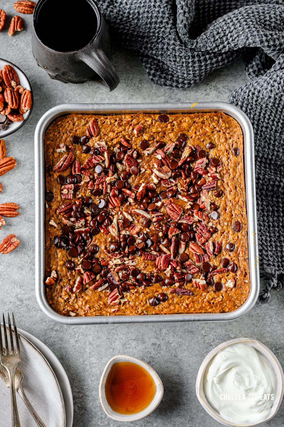 Baked Pumpkin Oatmeal in a baking pan and topped with pecans and chocolate chips, with Greek yogurt, maple syrup, and plates alongside.