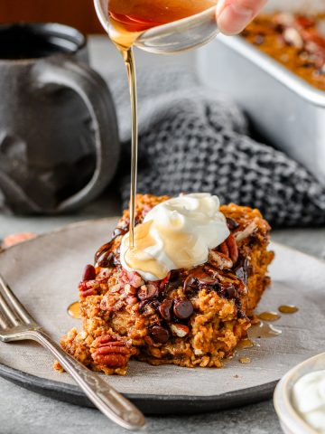 Serving of gluten free pumpkin baked oatmeal topped with Greek yogurt and drizzled with maple syrup on a ceramic plate with a cup of coffee in the background.