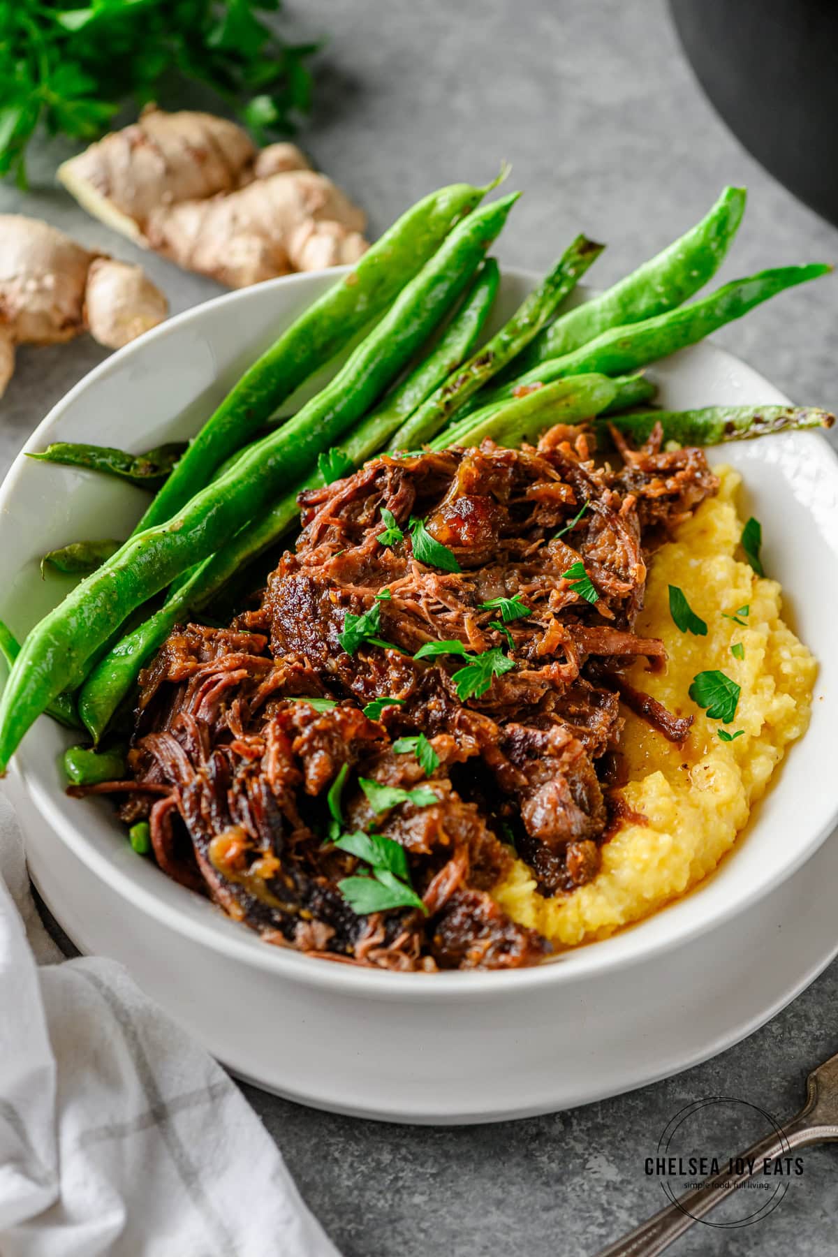 Shredded Braised Miso Ginger Chuck Roast served with polenta and sautéed green beans in a white bowl with ginger and linens in the background.