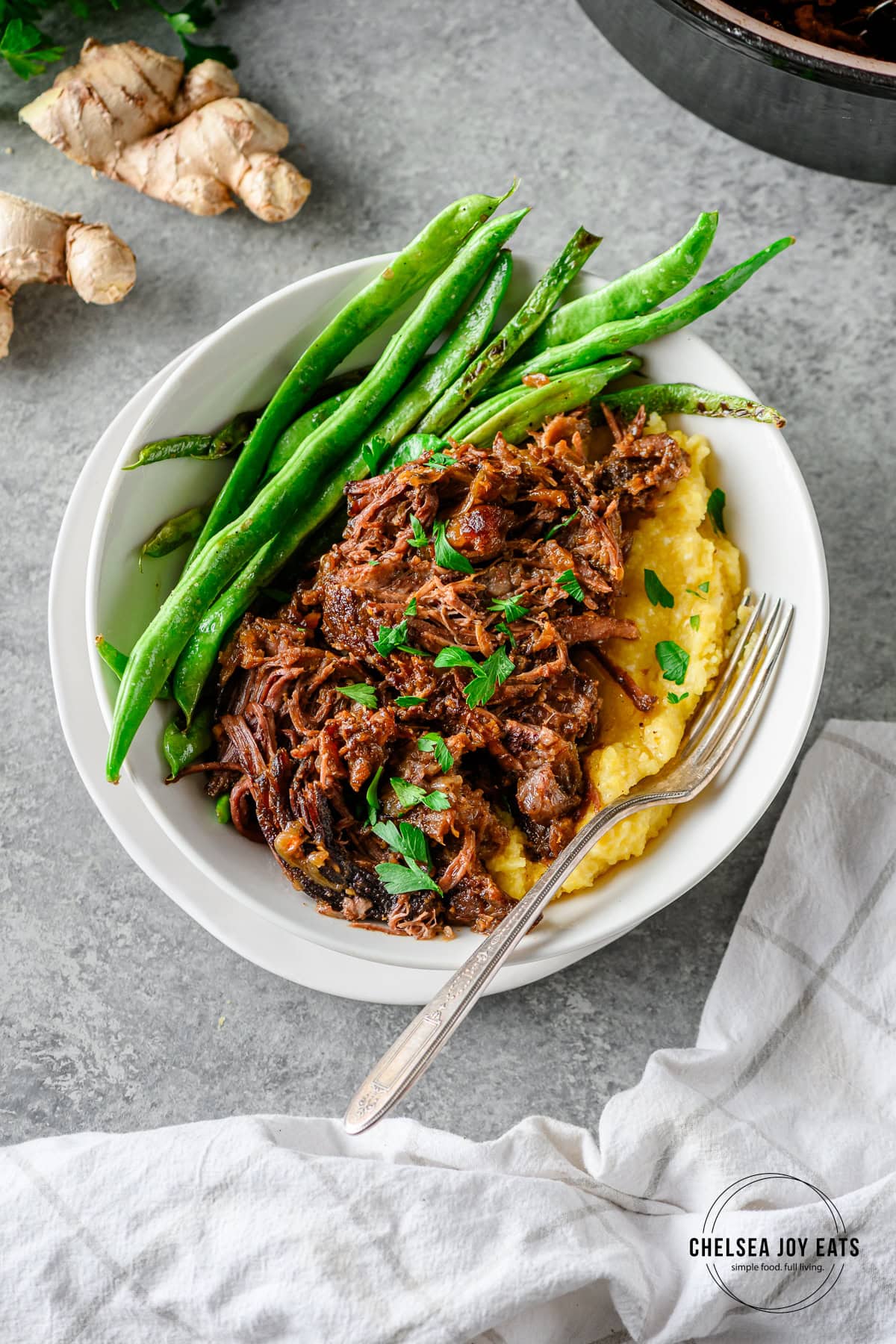Shredded Braised Miso Ginger Chuck Roast served with polenta and sautéed green beans in a white bowl with a fork.