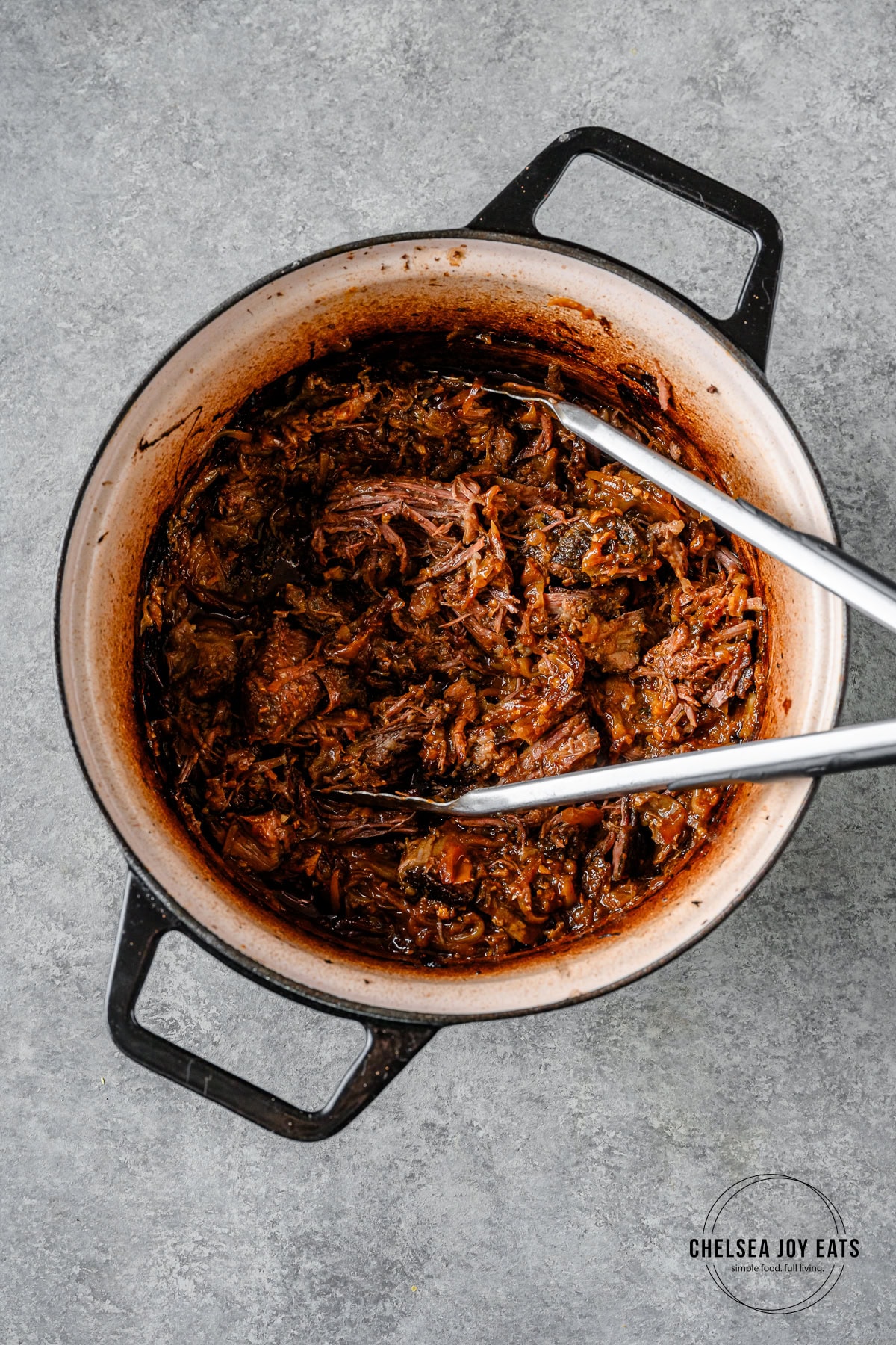 Shredded braised miso ginger chuck roast in a Dutch oven with kitchen tongs.