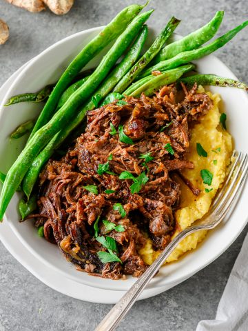 Shredded braised chuck roast served over polenta with sautéed green beans with a fork alongside.