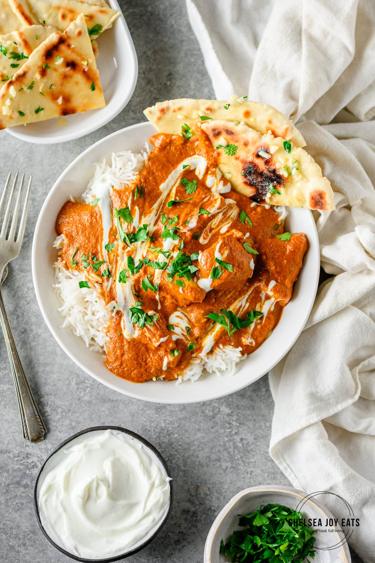 Bowl of chicken tikka masala with rice and gluten free flatbread in a white ceramic bowl on a gray background, with forks and linens besides.