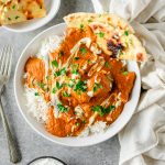 Bowl of chicken tikka masala with rice and gluten free flatbread in a white ceramic bowl on a gray background, with forks and linens besides.