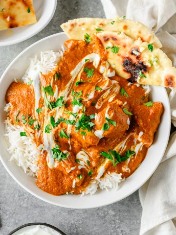 Bowl of chicken tikka masala with rice and gluten free flatbread in a white ceramic bowl on a gray background, with forks and linens besides.
